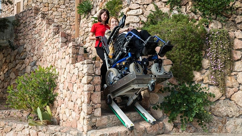 an elderly disabled person in a wheelchair is transported up an outdoor stone staircase with the electric wheelchair stair crawler